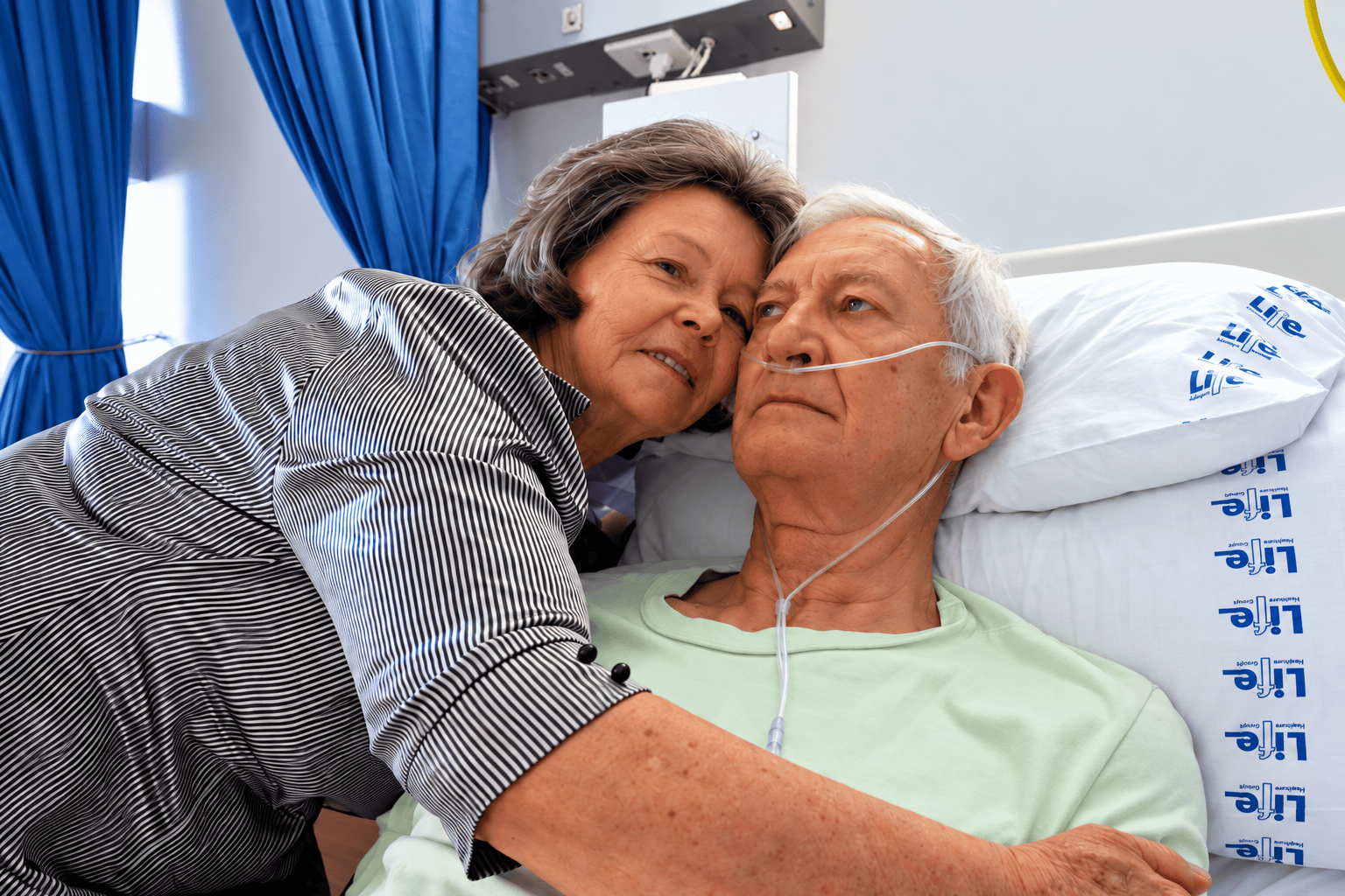 Older man resting in a hospital bed with oxygen support while a woman embraces him.